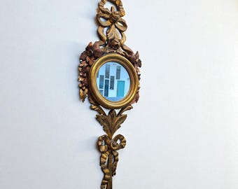 May include: Ornate, antique wall mirror with a round, gold-framed mirror. The mirror is surrounded by detailed, carved gold-toned embellishments, including flowers, leaves, and ribbons. The mirror is mounted on a white wall.