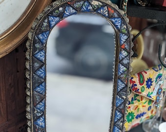 Puede incluir: Un espejo arqueado con un borde de mosaico con azulejos triangulares azules, negros y blancos. El marco tiene un acabado plateado y un borde texturizado. El espejo se exhibe con otros objetos decorativos.