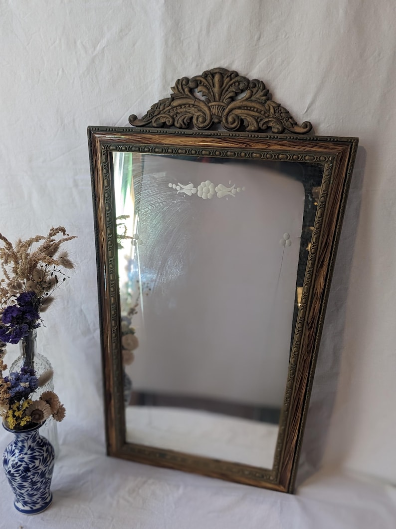 May include: A vintage wood framed mirror with a decorative carved top and etched floral design. The mirror has a distressed wood finish and a beveled edge.
