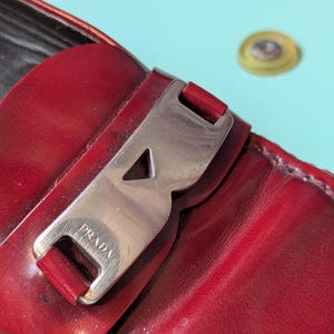 May include: Close-up of a red leather loafer with a silver-tone buckle. The buckle is engraved with the brand name "PRADA". The leather has a glossy finish. The shoe is on a light blue surface.