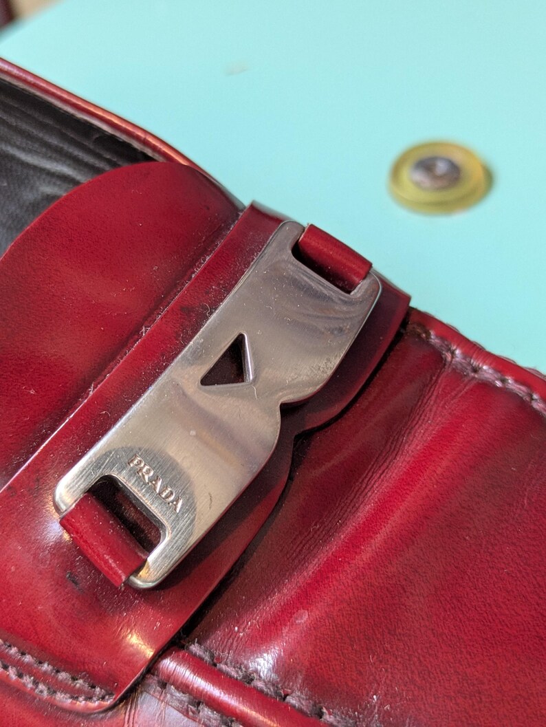 May include: Close-up of a red leather loafer with a silver-tone buckle. The buckle is engraved with the brand name "PRADA". The leather has a glossy finish. The shoe is on a light blue surface.