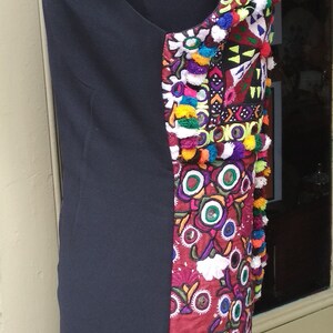 May include: A black sleeveless dress with a colorful embroidered panel on the right side. The embroidery features a variety of colors, including red, green, blue, and yellow. The embroidery also includes small mirrors and pom-poms.
