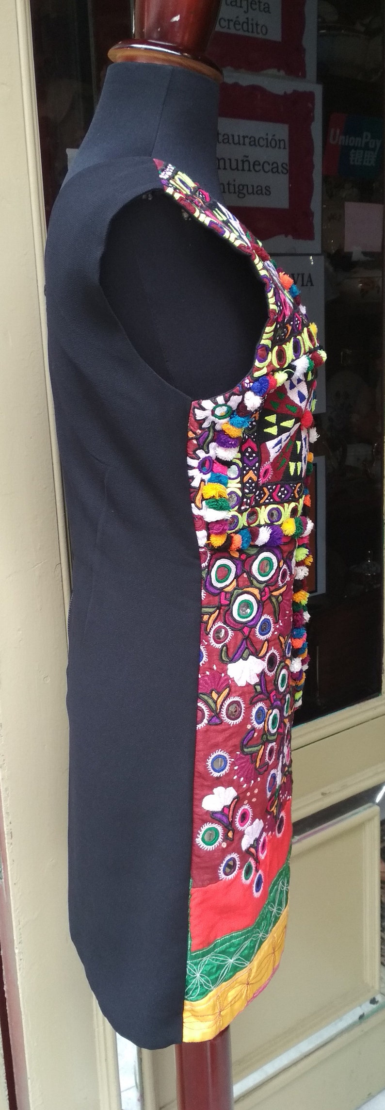 May include: A black sleeveless dress with a colorful embroidered panel on the right side. The embroidery features a variety of colors, including red, green, blue, and yellow. The embroidery also includes small mirrors and pom-poms.