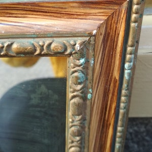May include: A gold-toned picture frame with a wood-grain finish and a decorative border. The frame has a green patina on the edges.