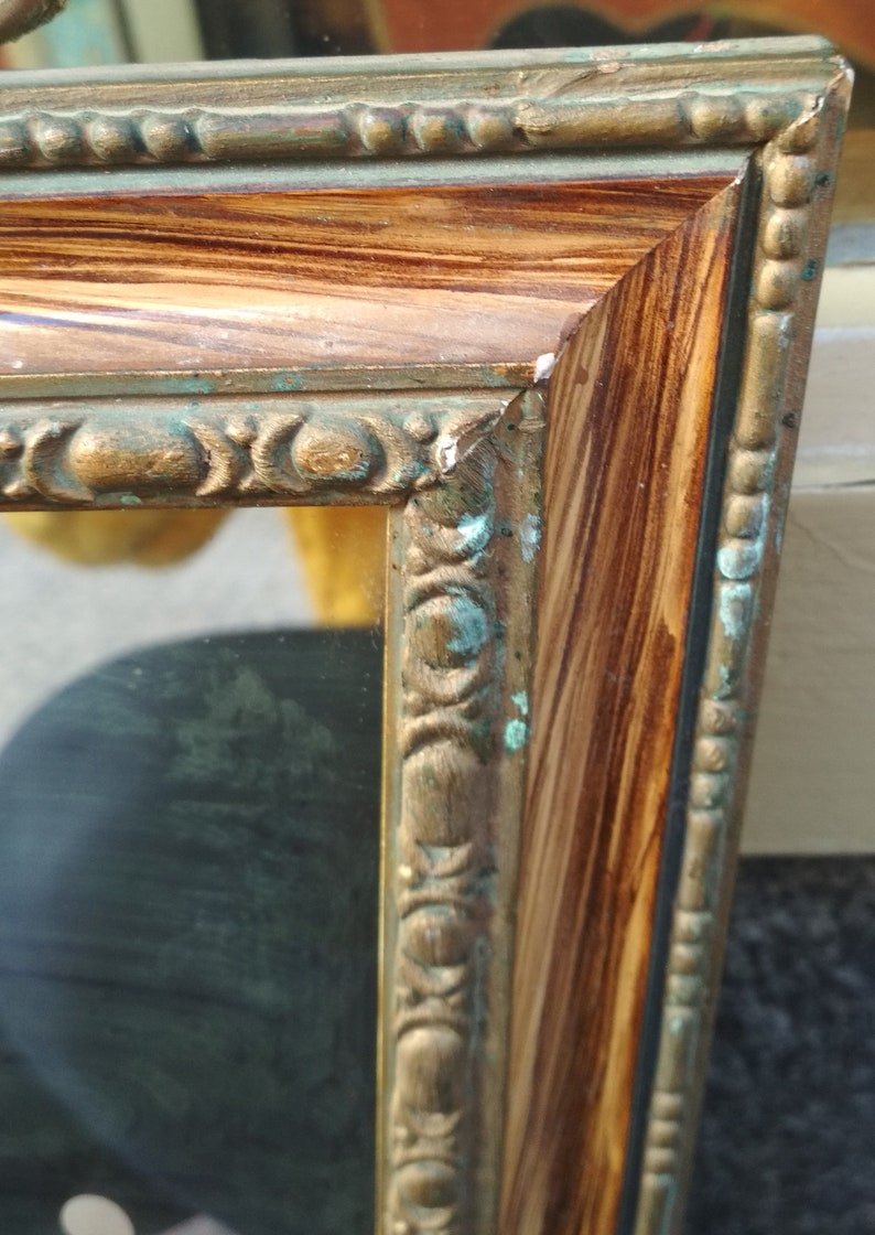 May include: A gold-toned picture frame with a wood-grain finish and a decorative border. The frame has a green patina on the edges.