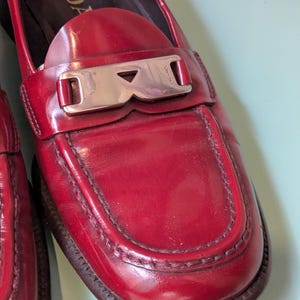 May include: Red leather loafers with a silver-tone buckle. The shoes have a classic design with a rounded toe and a stitched detail around the vamp. The brand name "PRADA" is visible on the buckle.