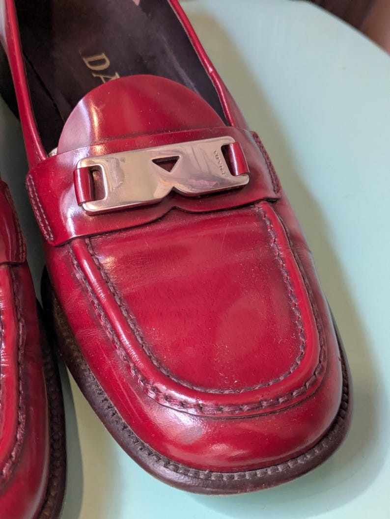 May include: Red leather loafers with a silver-tone buckle. The shoes have a classic design with a rounded toe and a stitched detail around the vamp. The brand name "PRADA" is visible on the buckle.