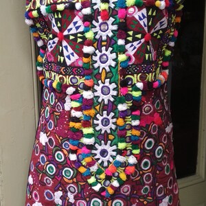 May include: A colorful embroidered vest with a V-neckline and a red, green, and yellow trim. The vest is decorated with pom-poms, mirrors, and intricate stitching.