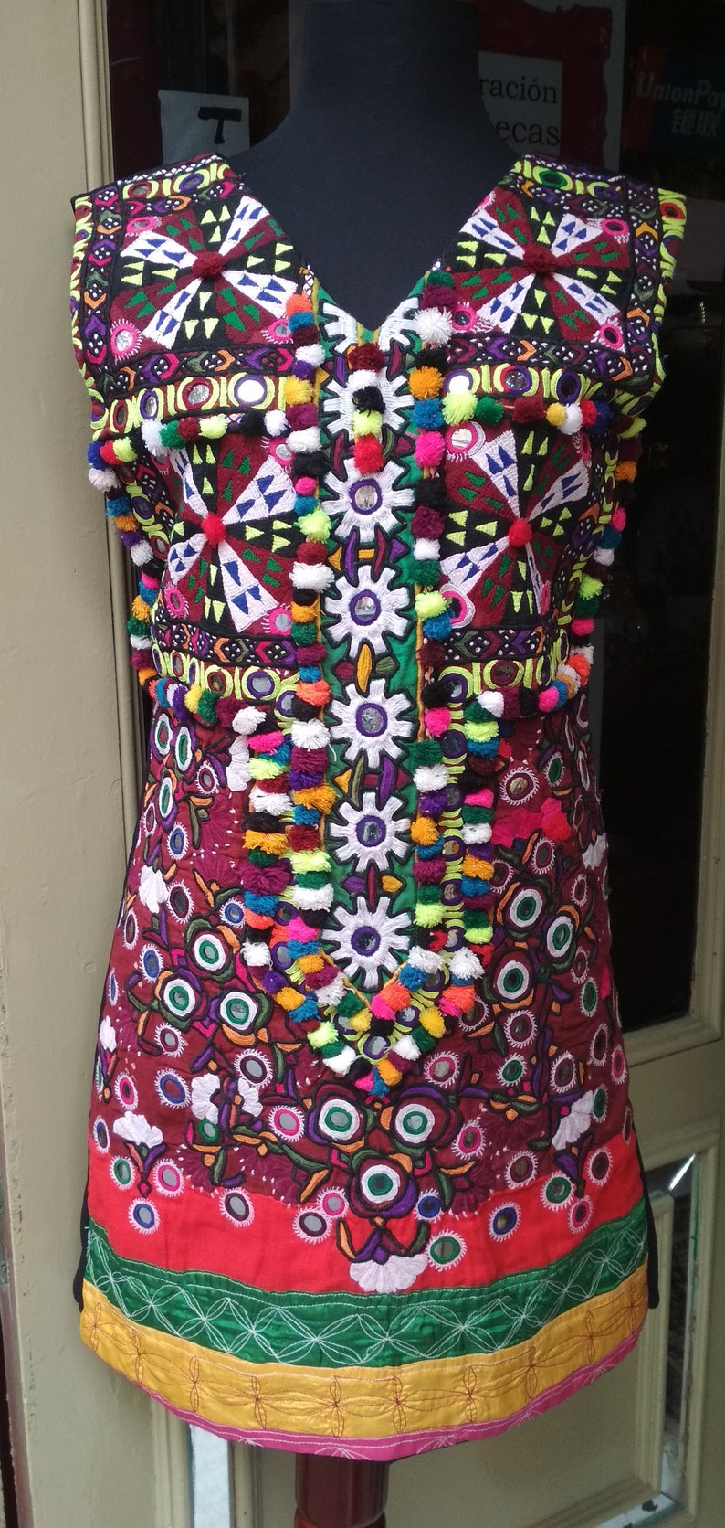 May include: A colorful embroidered vest with a V-neckline and a red, green, and yellow trim. The vest is decorated with pom-poms, mirrors, and intricate stitching.
