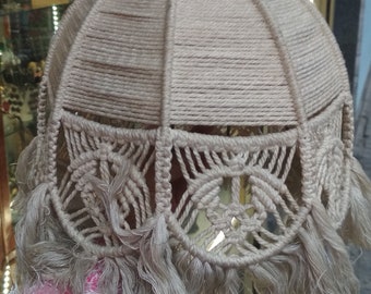 Puede incluir: Una pantalla de lámpara de macramé beige con la parte superior redonda y un borde de flecos. La pantalla de lámpara está hecha de cuerda tejida y tiene un patrón delicado e intrincado.