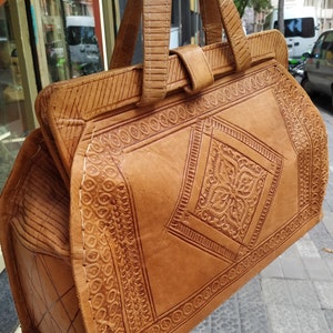 Puede incluir: Un bolso de mano de cuero marr&oacute;n con intrincados dise&ntilde;os en relieve. El bolso tiene dos asas superiores y un cierre de solapa.