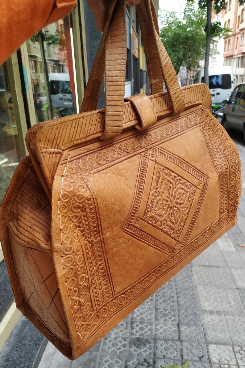 Puede incluir: Un bolso de mano de cuero marr&oacute;n con intrincados dise&ntilde;os en relieve. El bolso tiene dos asas superiores y un cierre de solapa.