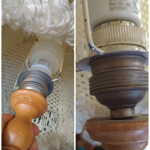 Op de afbeelding: Close-up van een lampvoet met een witte lampenkap en een gloeilamp. De lampvoet is gemaakt van hout en heeft een metalen fitting. De lampenkap is gemaakt van witte stof met een franje.