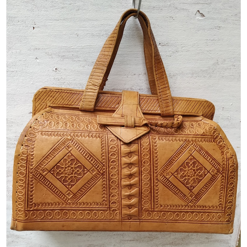 Puede incluir: Un bolso de mano de cuero marr&oacute;n con intrincados patrones en relieve y un asa superior. El bolso tiene un cierre de solapa con una hebilla decorativa.