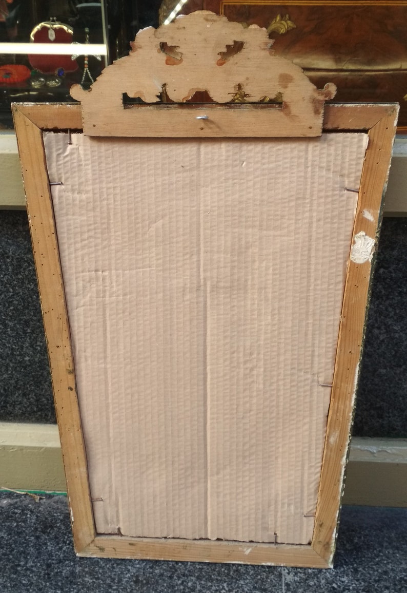 May include: A vintage wooden picture frame with a gold-painted finish. The frame has a decorative top with a carved design. The back of the frame is made of cardboard.