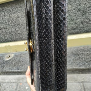 Puede incluir: Bolso de mano de cuero negro con un patr&oacute;n texturizado de piel de serpiente. El bolso tiene un cierre dorado y una solapa de cierre.
