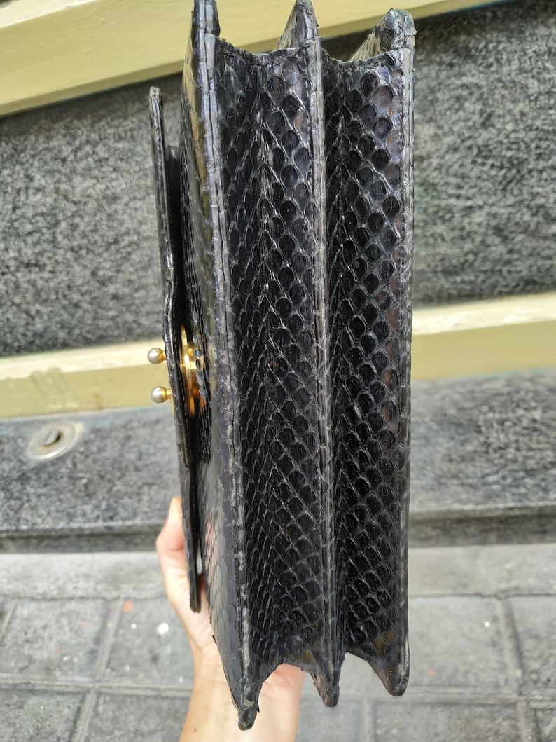 Puede incluir: Bolso de mano de cuero negro con un patr&oacute;n texturizado de piel de serpiente. El bolso tiene un cierre dorado y una solapa de cierre.
