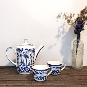 May include: A white ceramic teapot and two cups with blue and white floral designs. The teapot features a bird design on the front. The cups have a floral design on the front. The teapot and cups are sitting on a wooden table.