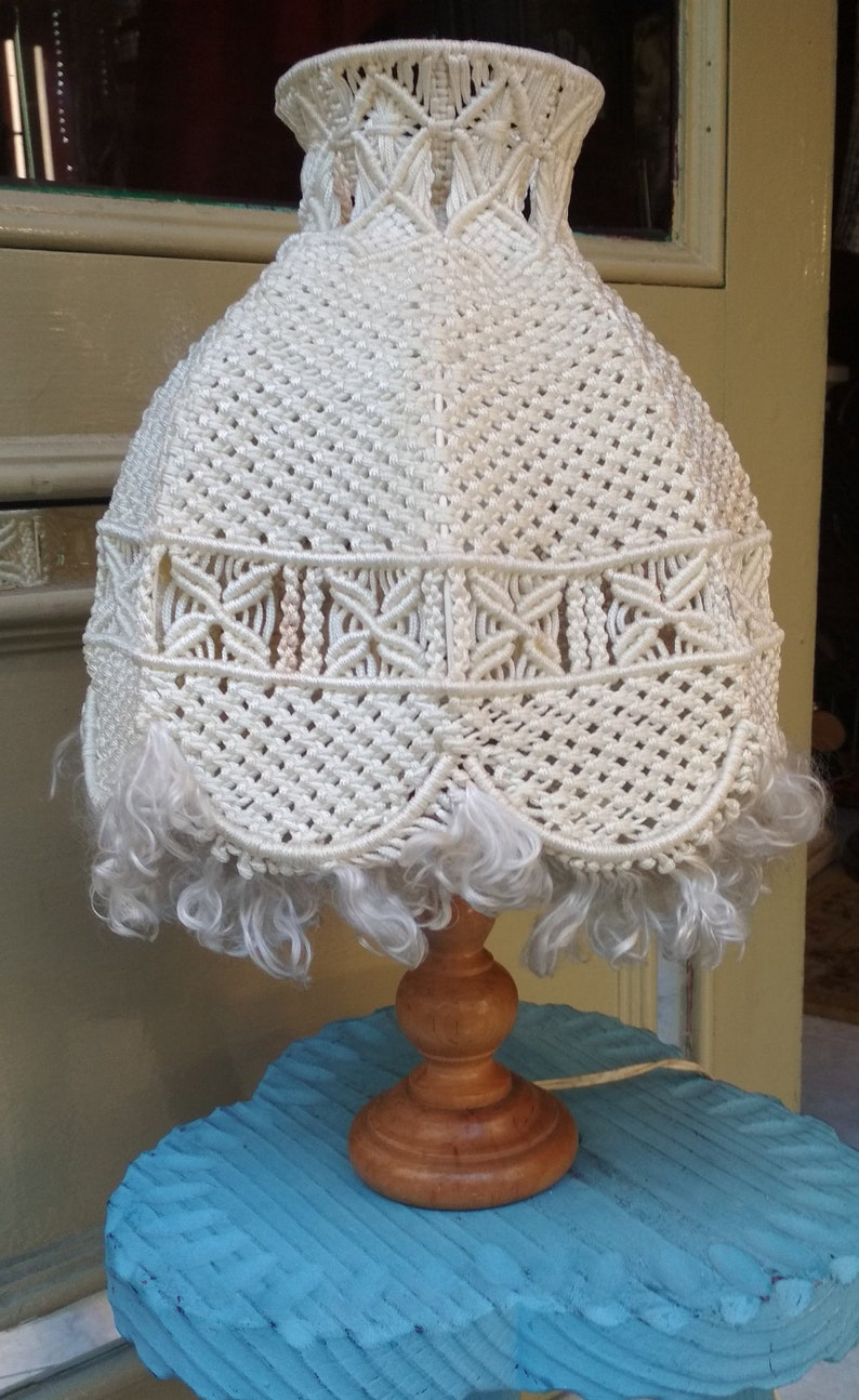 Op de afbeelding: Een witte macram&eacute; lampenkap met een franje rand op een blauwe houten basis. De lampenkap heeft een uniek ontwerp met ingewikkelde knopen en een gegolfde rand.