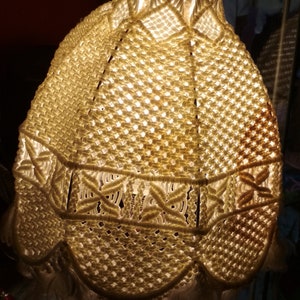 Op de afbeelding: Een witte macram&eacute; lampenkap met een houten voet. De lampenkap is gemaakt van een delicaat, ingewikkeld patroon van knopen.