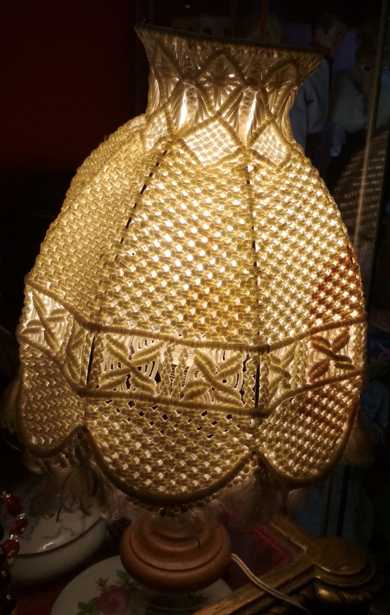 Op de afbeelding: Een witte macram&eacute; lampenkap met een houten voet. De lampenkap is gemaakt van een delicaat, ingewikkeld patroon van knopen.