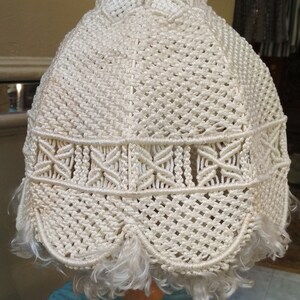 Op de afbeelding: Een witte macram&eacute; lampenkap met een franje rand op een houten voet. De lampenkap is gemaakt van een geweven koord en heeft een delicaat, ingewikkeld ontwerp.