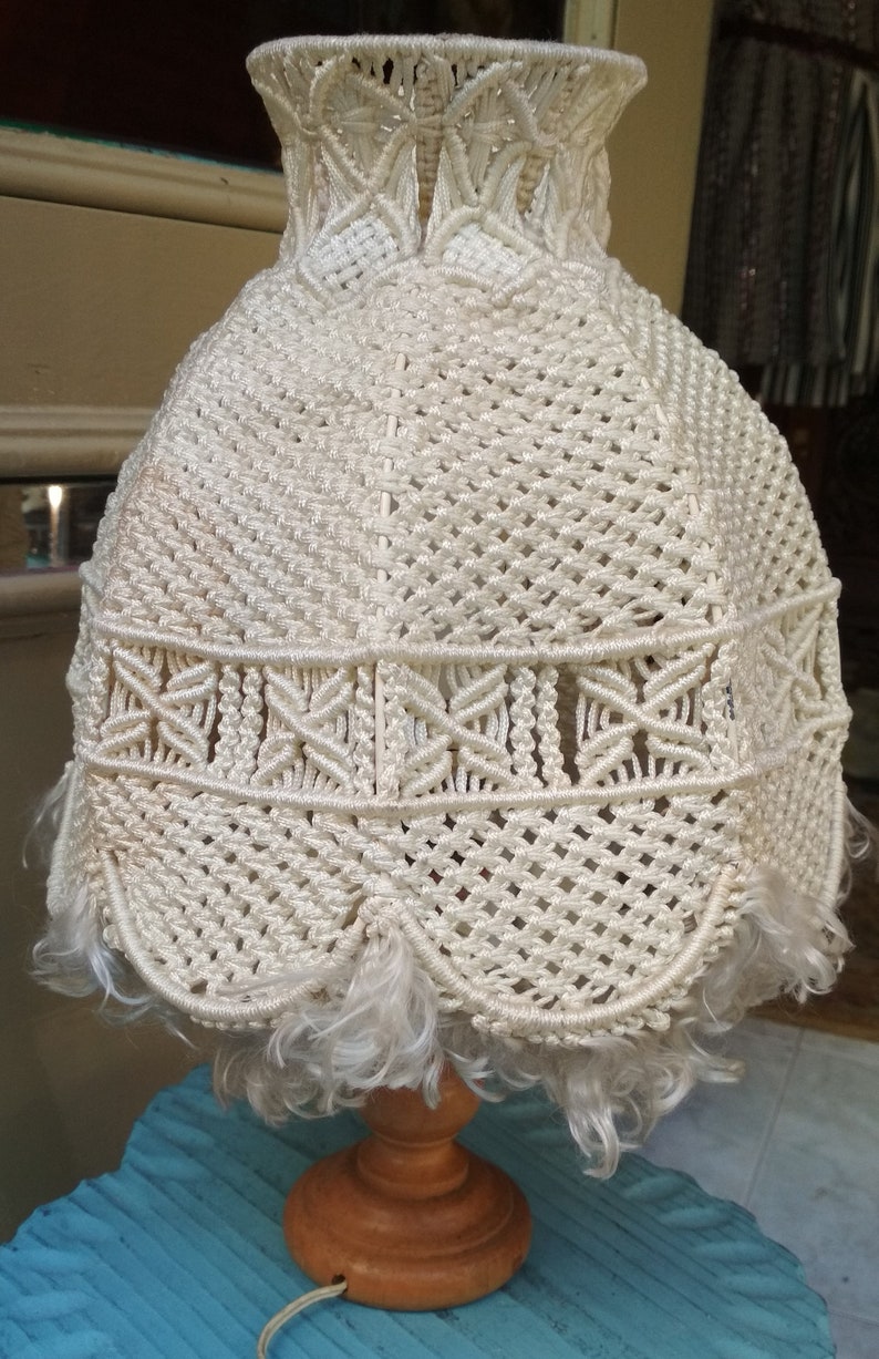 Op de afbeelding: Een witte macram&eacute; lampenkap met een franje rand op een houten voet. De lampenkap is gemaakt van een geweven koord en heeft een delicaat, ingewikkeld ontwerp.
