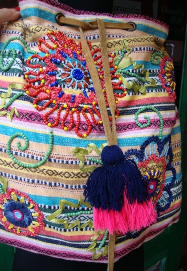 May include: A colorful striped drawstring bag with embroidered flowers and beads. The bag has a brown leather strap and a pink and blue tassel.