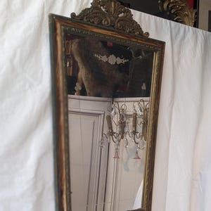 May include: A vintage ornate wood framed mirror with a gold finish and a beveled glass mirror. The mirror has a decorative top with a floral design.