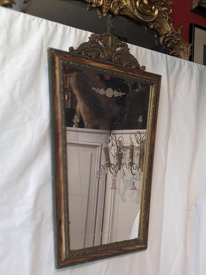 May include: A vintage ornate wood framed mirror with a gold finish and a beveled glass mirror. The mirror has a decorative top with a floral design.