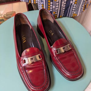 May include: A pair of red leather loafers with silver-tone hardware. The shoes have a classic penny loafer design with a rounded toe and a low heel. The brand name "PRADA" is visible on the insole.