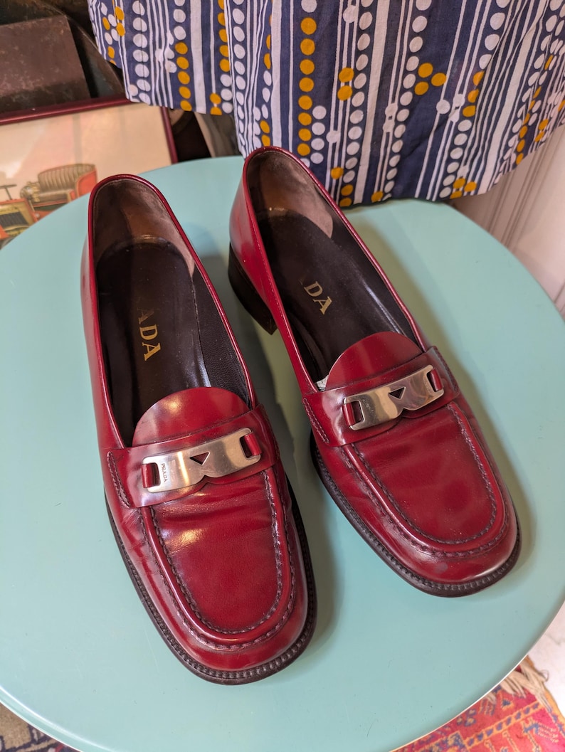 May include: A pair of red leather loafers with silver-tone hardware. The shoes have a classic penny loafer design with a rounded toe and a low heel. The brand name "PRADA" is visible on the insole.