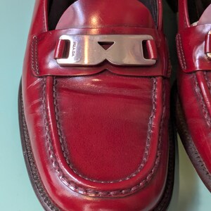 May include: Close-up of a pair of red leather loafers. The shoes have a silver-tone metal buckle with the brand name "Prada" on it. The loafers have a stitched design around the toe and a dark brown sole.