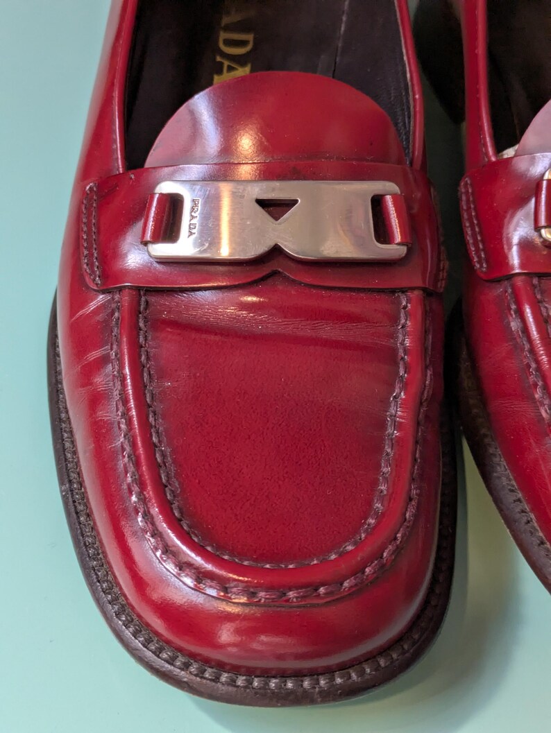 May include: Close-up of a pair of red leather loafers. The shoes have a silver-tone metal buckle with the brand name "Prada" on it. The loafers have a stitched design around the toe and a dark brown sole.