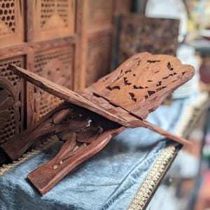 May include: A wooden book stand with intricate carved floral and geometric designs. The stand is made of dark brown wood and has a hinged design for easy storage. The stand is designed to hold a book open for reading.