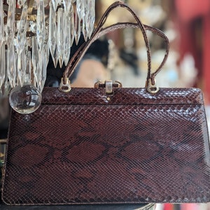 Op de afbeelding: Een vintage bruine handtas van slangenleer met een gouden sluiting en twee handvatten.