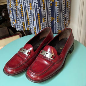 May include: A pair of red leather loafers with silver-tone metal accents. The shoes have a classic design with a low heel and a decorative strap across the top. The loafers are displayed on a light blue surface.