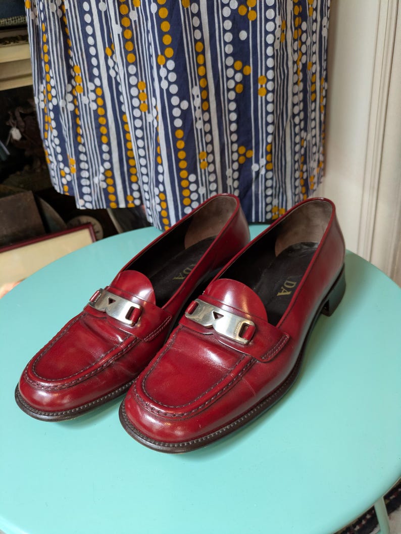 May include: A pair of red leather loafers with silver-tone metal accents. The shoes have a classic design with a low heel and a decorative strap across the top. The loafers are displayed on a light blue surface.