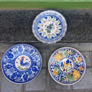 May include: Three decorative ceramic plates with hand-painted designs. The plates feature blue, white, and yellow floral and rooster motifs. The plates are arranged on a dark gray surface.