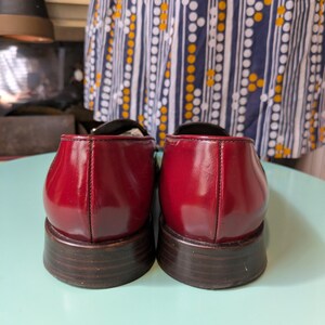May include: Pair of glossy, burgundy leather shoes with a low, stacked heel. The shoes have a classic design with a rounded toe and a subtle seam detail down the back. The heels are approximately 1 inch (2.5 cm) high.