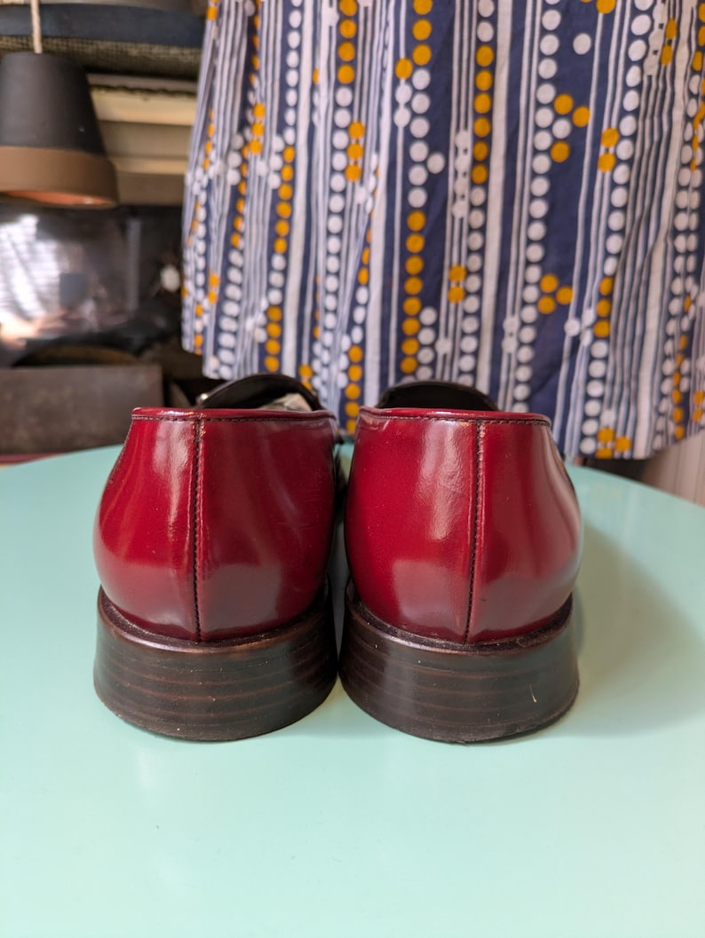 May include: Pair of glossy, burgundy leather shoes with a low, stacked heel. The shoes have a classic design with a rounded toe and a subtle seam detail down the back. The heels are approximately 1 inch (2.5 cm) high.