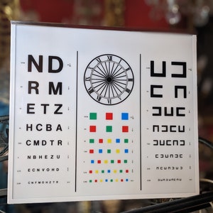 May include: A white eye chart with black letters and a clock face. The chart includes a colour vision test with rows of coloured squares.