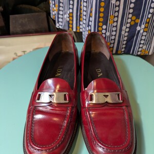 May include: A pair of red leather loafers with silver-tone hardware. The shoes have a classic design with a rounded toe and a low heel. The word "PRADA" is visible on the insole. The loafers are displayed on a light blue surface.
