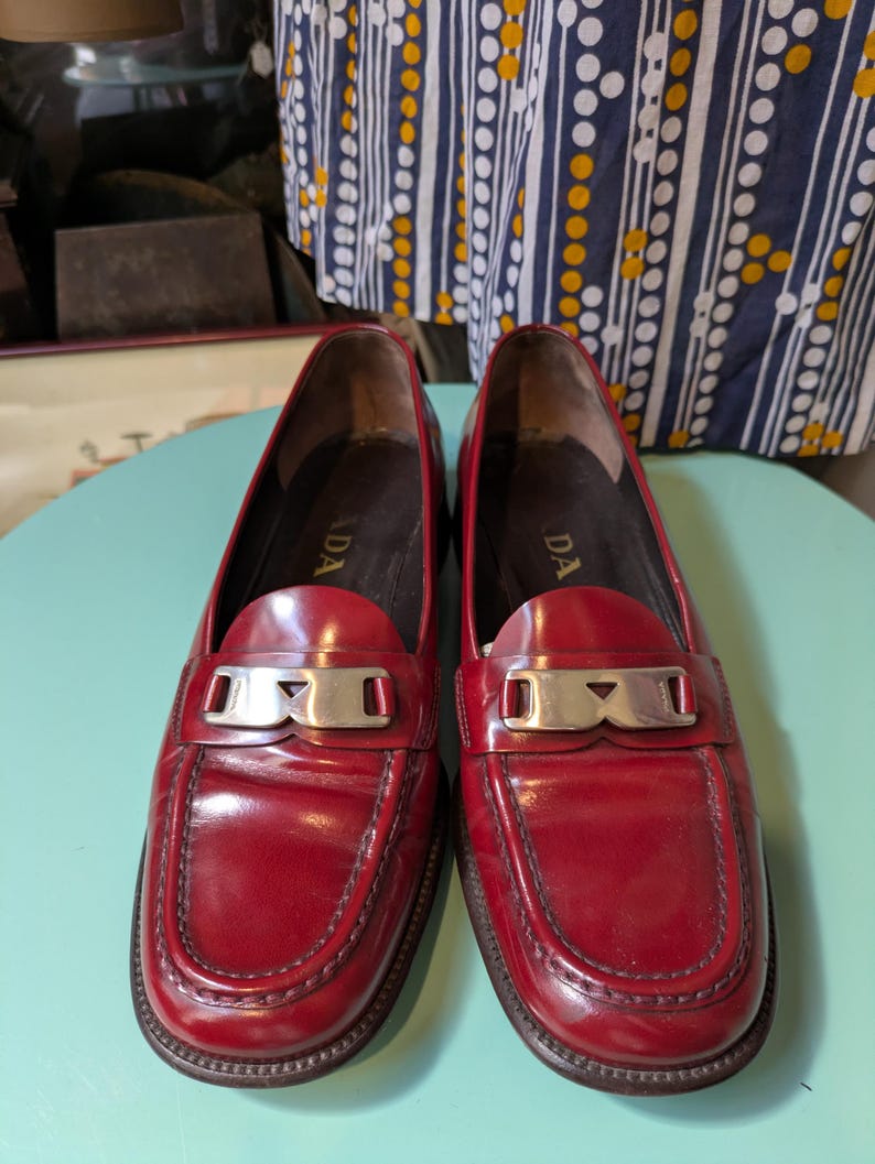 May include: A pair of red leather loafers with silver-tone hardware. The shoes have a classic design with a rounded toe and a low heel. The word "PRADA" is visible on the insole. The loafers are displayed on a light blue surface.