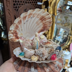May include: A decorative seashell sculpture featuring a small statue of a religious figure, surrounded by various seashells and colorful gems. The sculpture is held in a hand, showcasing its intricate details and craftsmanship. The sculpture is a unique piece of art.