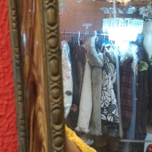 May include: A wooden framed mirror with an ornate gold trim. The mirror reflects a room with a chandelier and clothing hanging on a rack.