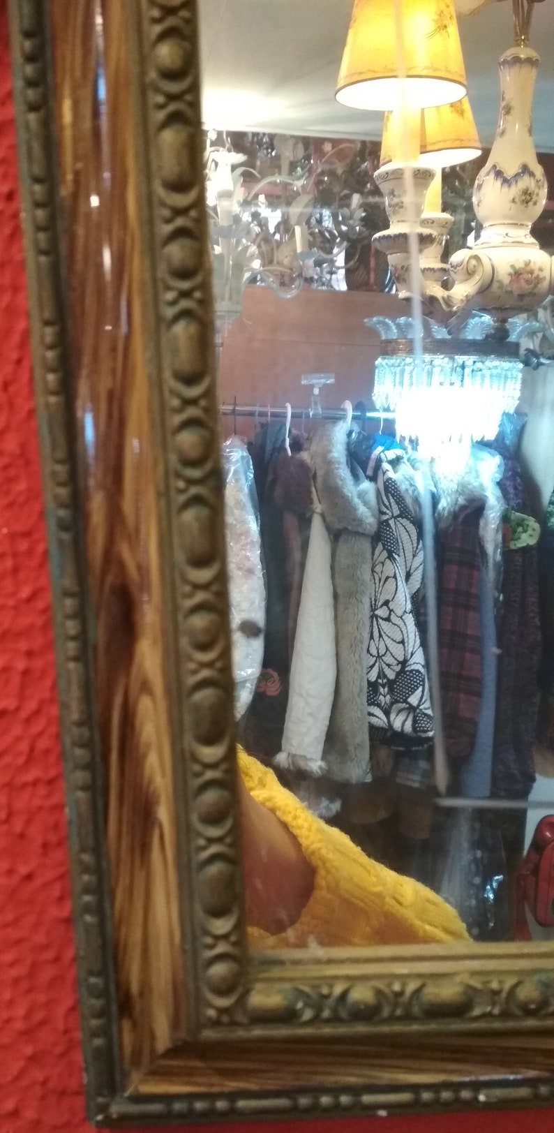 May include: A wooden framed mirror with an ornate gold trim. The mirror reflects a room with a chandelier and clothing hanging on a rack.