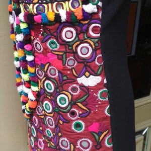 May include: A black dress with a colorful embroidered panel featuring floral designs, mirrors, and pom-poms. The embroidery is in shades of red, green, blue, yellow, and white.