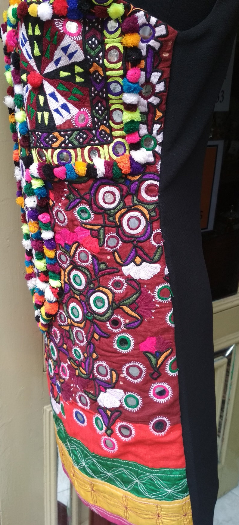 May include: A black dress with a colorful embroidered panel featuring floral designs, mirrors, and pom-poms. The embroidery is in shades of red, green, blue, yellow, and white.