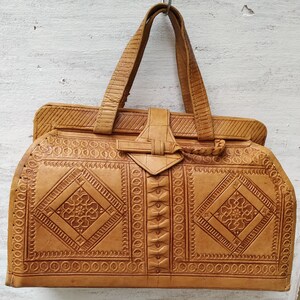 Puede incluir: Un bolso de mano de cuero marr&oacute;n con intrincados patrones en relieve. El bolso tiene un asa superior y un cierre de solapa con una hebilla decorativa.